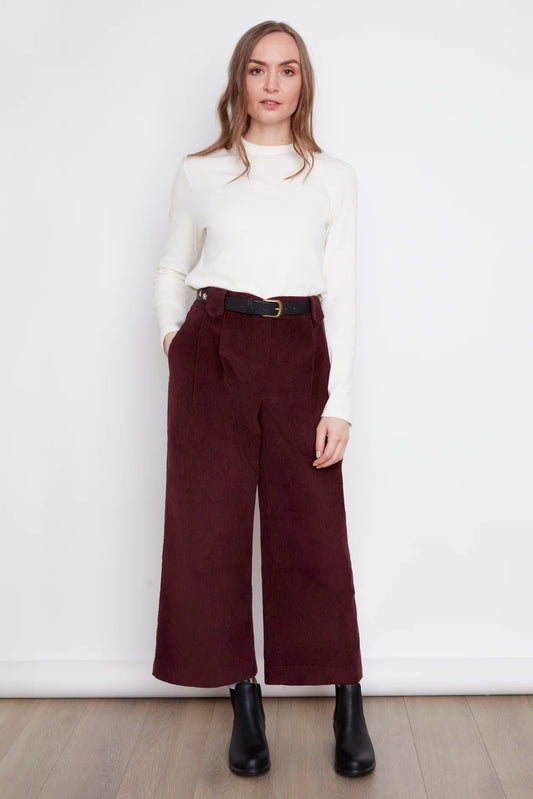 A woman wearing the Idadora Pants by Julei in Bordeaux, wide-leg corduroy pants. She is wearing them with a white sweater and standing against a white background