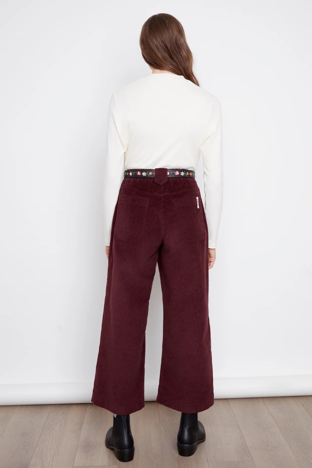 Back view of a woman wearing the Idadora Pants by Julei in Bordeaux, wide-leg corduroy pants. She is wearing them with a white sweater and standing against a white background