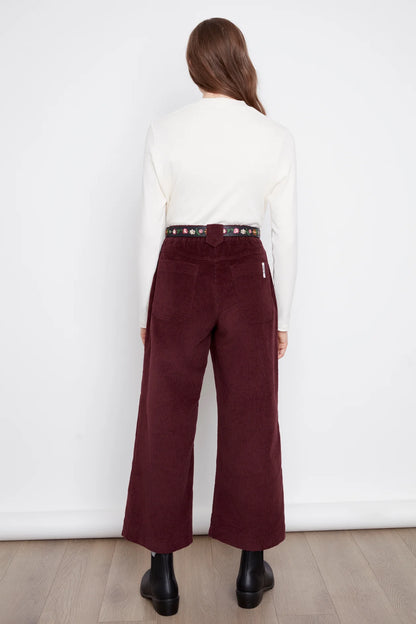 Back view of a woman wearing the Idadora Pants by Julei in Bordeaux, wide-leg corduroy pants. She is wearing them with a white sweater and standing against a white background