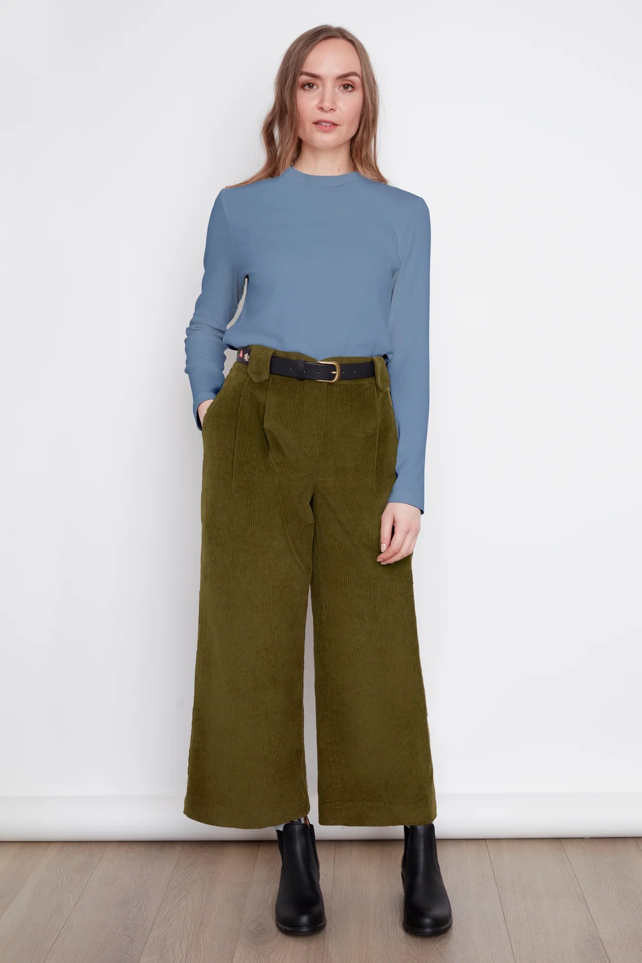A woman wearing the Idadora Pants by Julei in Moss, wide-leg corduroy pants. She is wearing them with a blue sweater and standing against a white background