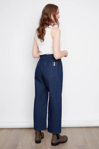 Bak view of a woman wearing the Alice Straight Leg Jeans by Julei in Dark Denim, with an elastic back waistband, seams at the front and back of the legs, and pointed belt loops. She is wearing them with a white tank and standing in front of a white background. 