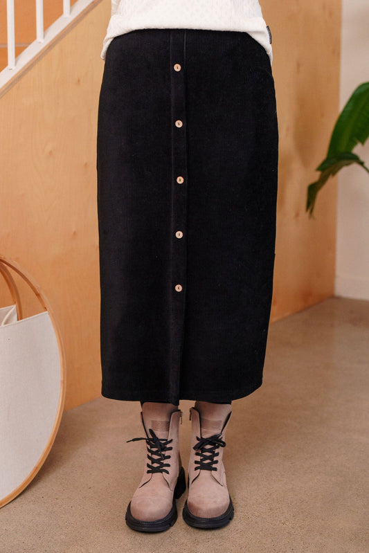 Waist down view of a woman wearing the Pruche Skirt by Rien ne se Perd, a 7/8 length Black corduroy skirt with an elastic waist, five coconut buttons up the front, and a slit at the back. She is wearing it with a white top and standing indoors. 