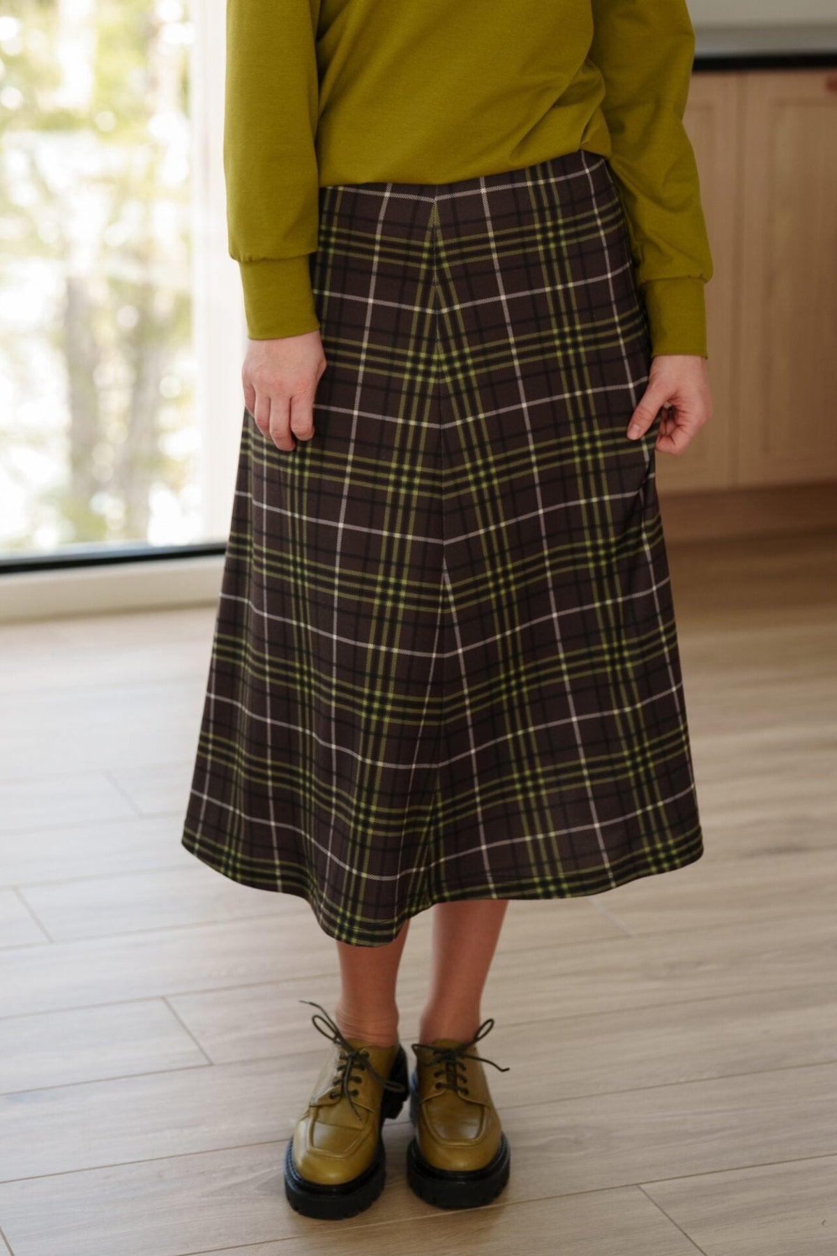 Waist down view of a woman standing in a white living room wearing the Hudson Skirt by Rien ne se Perd in Chocolate and Pistachio Plaid and a green top. 