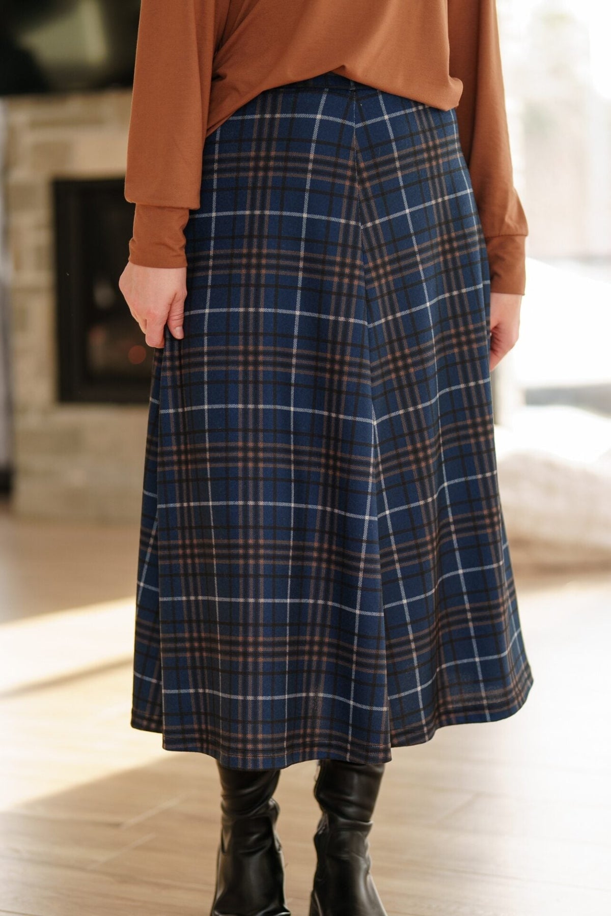 Waist down view of a woman standing in a white living room wearing the Hudson Skirt by Rien ne se Perd in Navy and Caramel  Plaid and a brown top. 