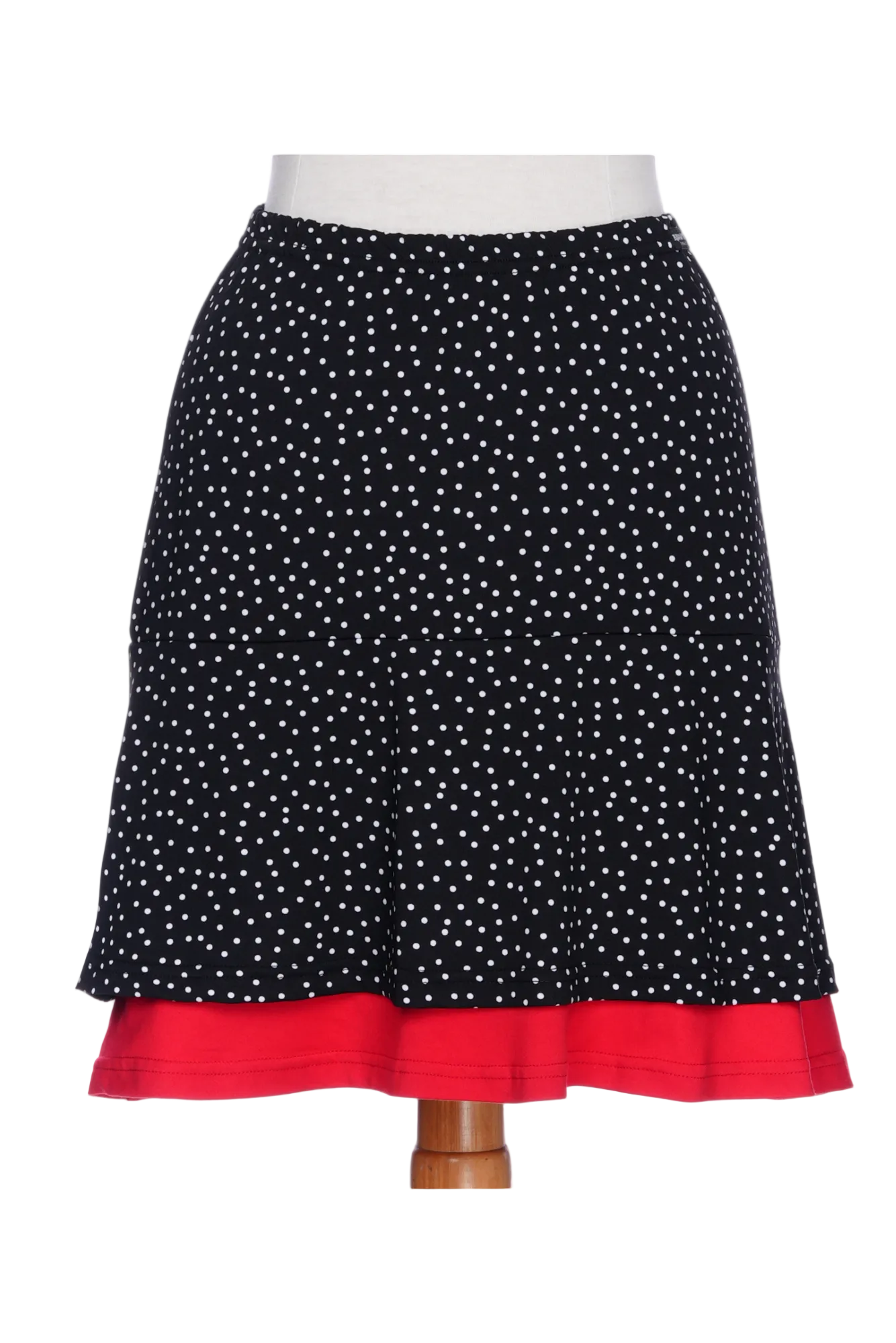 The Sambuca Skirt from Rien ne se Perd in black and white polka dots with a Red peekaboo ruffle is show on a mannequin against a white background. It has an elastic waist and an A-line shape that hits above the knee. 