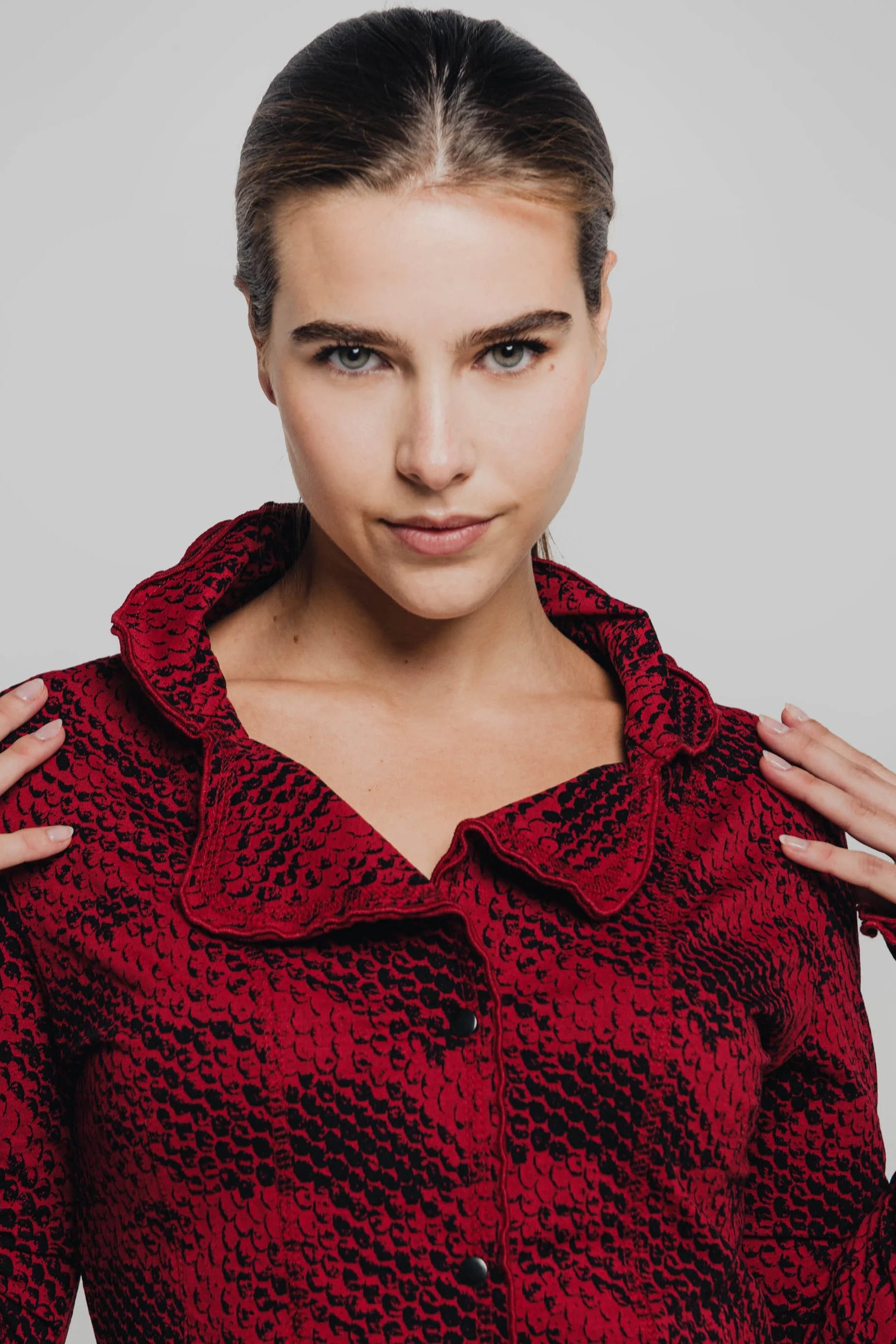 Waist up view of a woman wearing the Essential Jacket by Karkass in Red Snakeskin, a fitted jacket with a ruffled collar, button front, and frills at the hem and cuffs. She is standing in front of a grey background. 