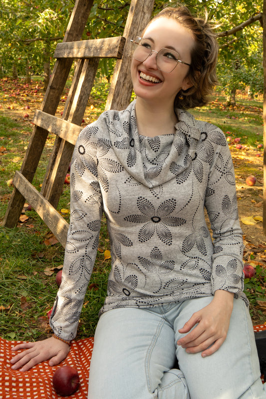 A woman wearing the Teddy Pearl Sweater by Karkass in Grey and Black, a cowl neck sweater with a floral pattern in black dots. She is sitting outdoors with trees and grass in the background.