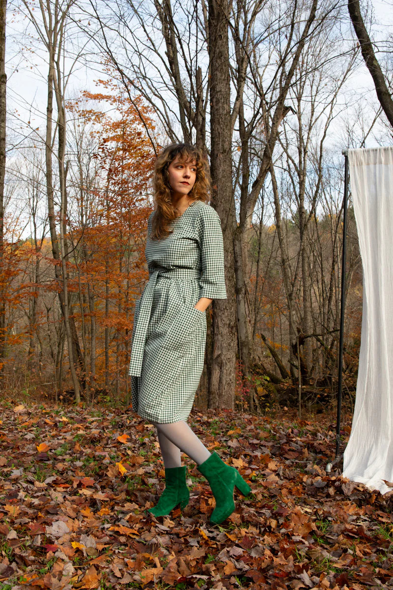 A woman wearing the Persea Dress by Kazak in Forest vichy print, a below-the-knee length dress with 3/4 sleeves, a removable belt, and large patch pockets. She is standing in a forest in beside a white sheet. 