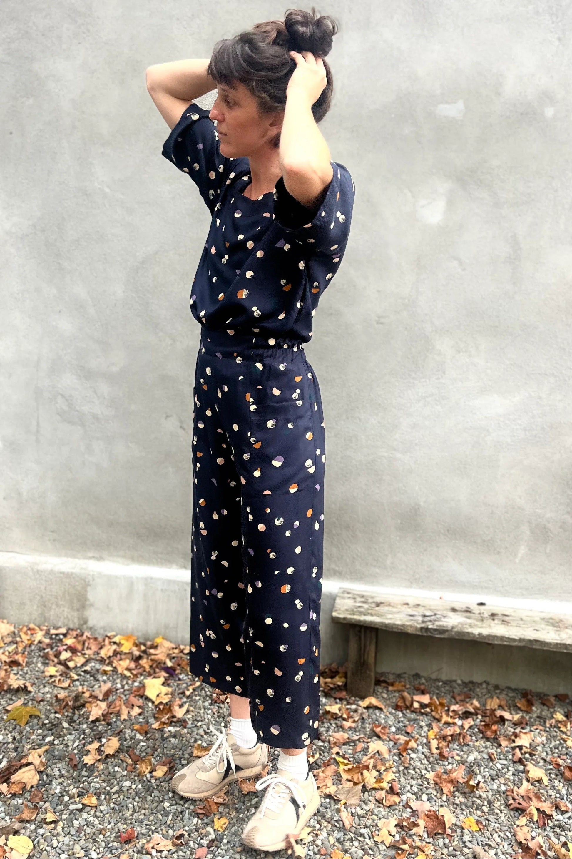 A woman wearing the Olea Pants by 
Kazak in Midnight Moons, wide-legged ankle-length pants with a flat-front elastic waist and large patch pockets. She is wearing them with the matching Abelia Top and standing outside by a grey wall. 