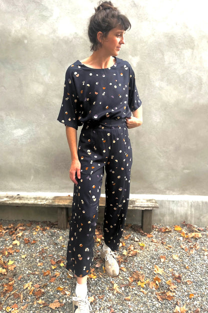 A woman wearing the Olea Pants by 
Kazak in Midnight Moons, wide-legged ankle-length pants with a flat-front elastic waist and large patch pockets. She is wearing them with the matching Abelia Top and standing outside by a grey wall. 