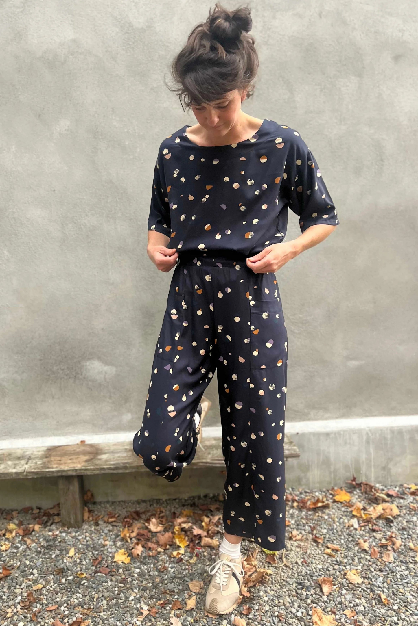 A woman wearing the Olea Pants by 
Kazak in Midnight Moons, wide-legged ankle-length pants with a flat-front elastic waist and large patch pockets. She is wearing them with the matching Abelia Top and standing outside by a grey wall. 