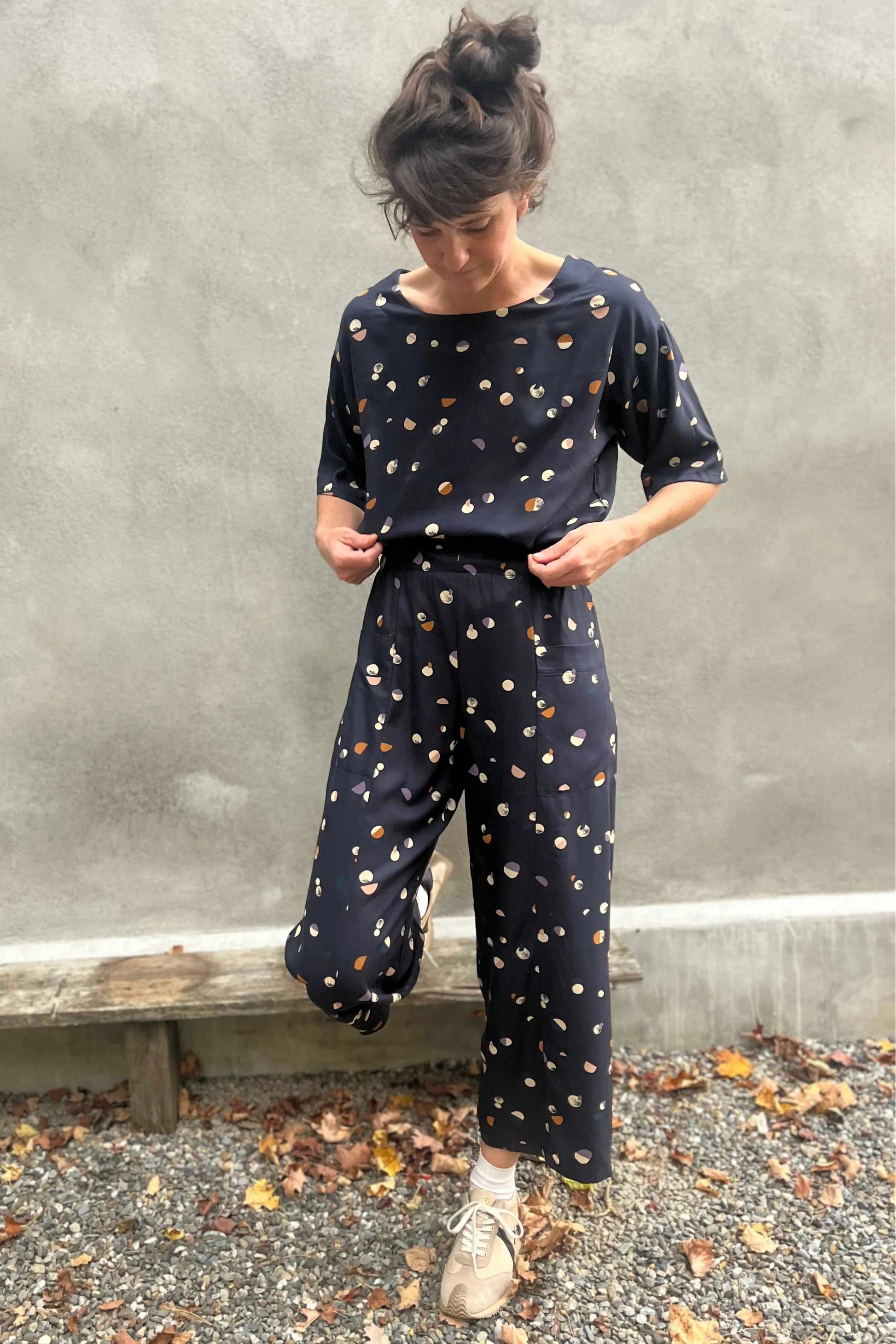 A woman wearing the Olea Pants by 
Kazak in Midnight Moons, wide-legged ankle-length pants with a flat-front elastic waist and large patch pockets. She is wearing them with the matching Abelia Top and standing outside by a grey wall. 