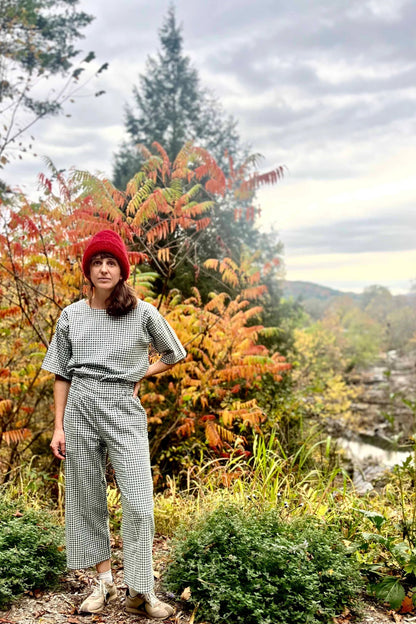 A woman wearing the Olea Pants by 
Kazak in Forest Vichy, wide-legged ankle-length pants with a flat-front elastic waist and large patch pockets. She is wearing them with the matching Abelia Top and standing in a forest.