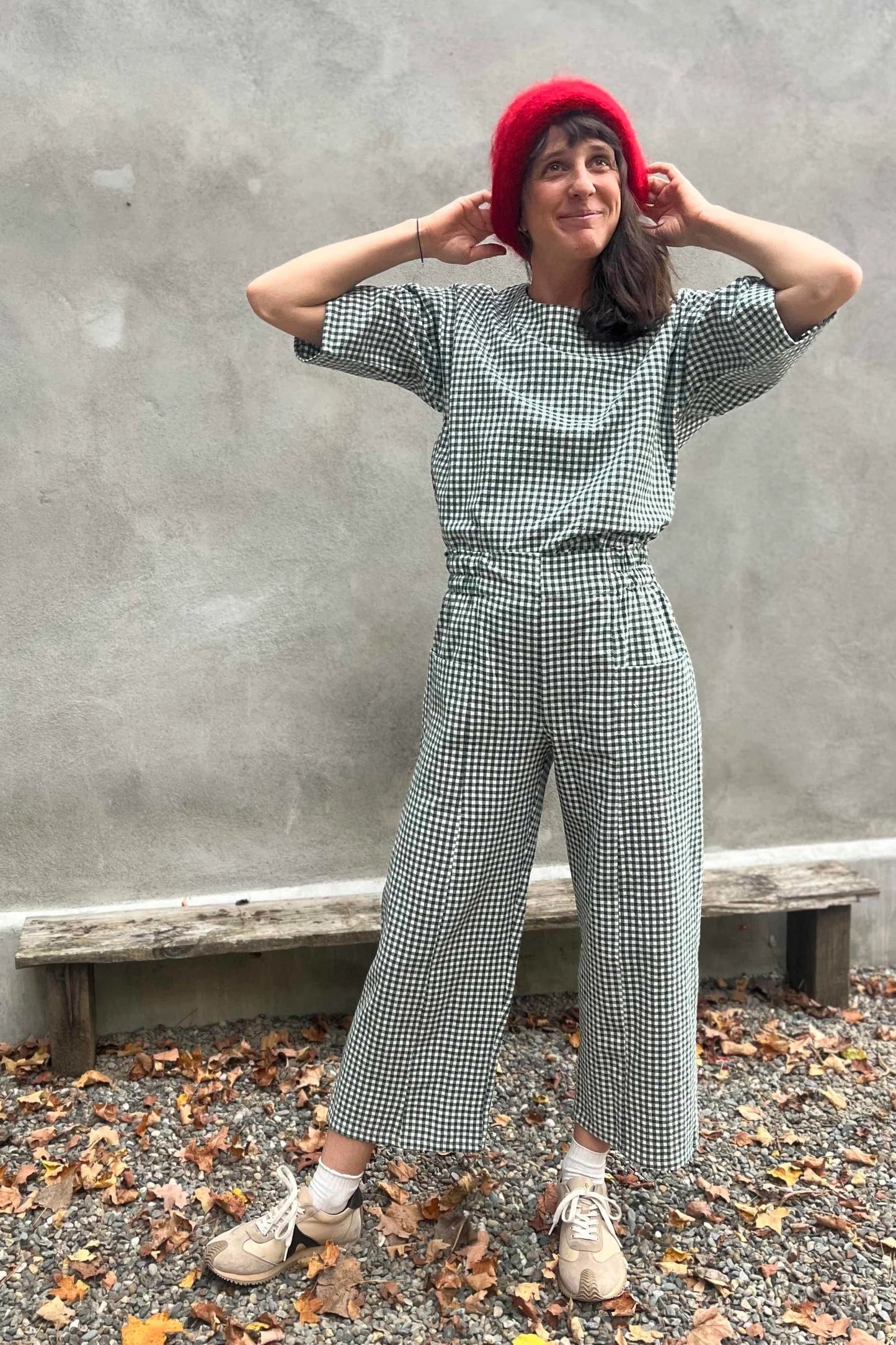 A woman wearing the Olea Pants by 
Kazak in Forest Vichy, wide-legged ankle-length pants with a flat-front elastic waist and large patch pockets. She is wearing them with the matching Abelia Top and standing outside by a grey wall. 
