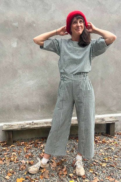 A woman wearing the Olea Pants by 
Kazak in Forest Vichy, wide-legged ankle-length pants with a flat-front elastic waist and large patch pockets. She is wearing them with the matching Abelia Top and standing outside by a grey wall. 
