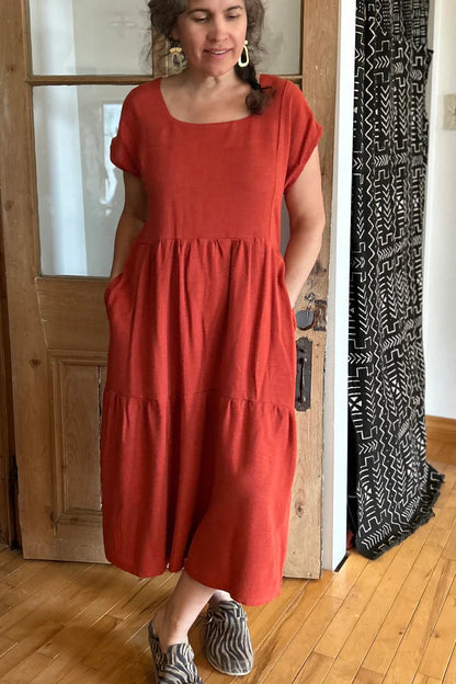 Woman wearing the Lavendula Dress by Kazak in Paprika, short sleeved square neck midi dress with three ruffles. She is standing in a room with wooden floors and a door.