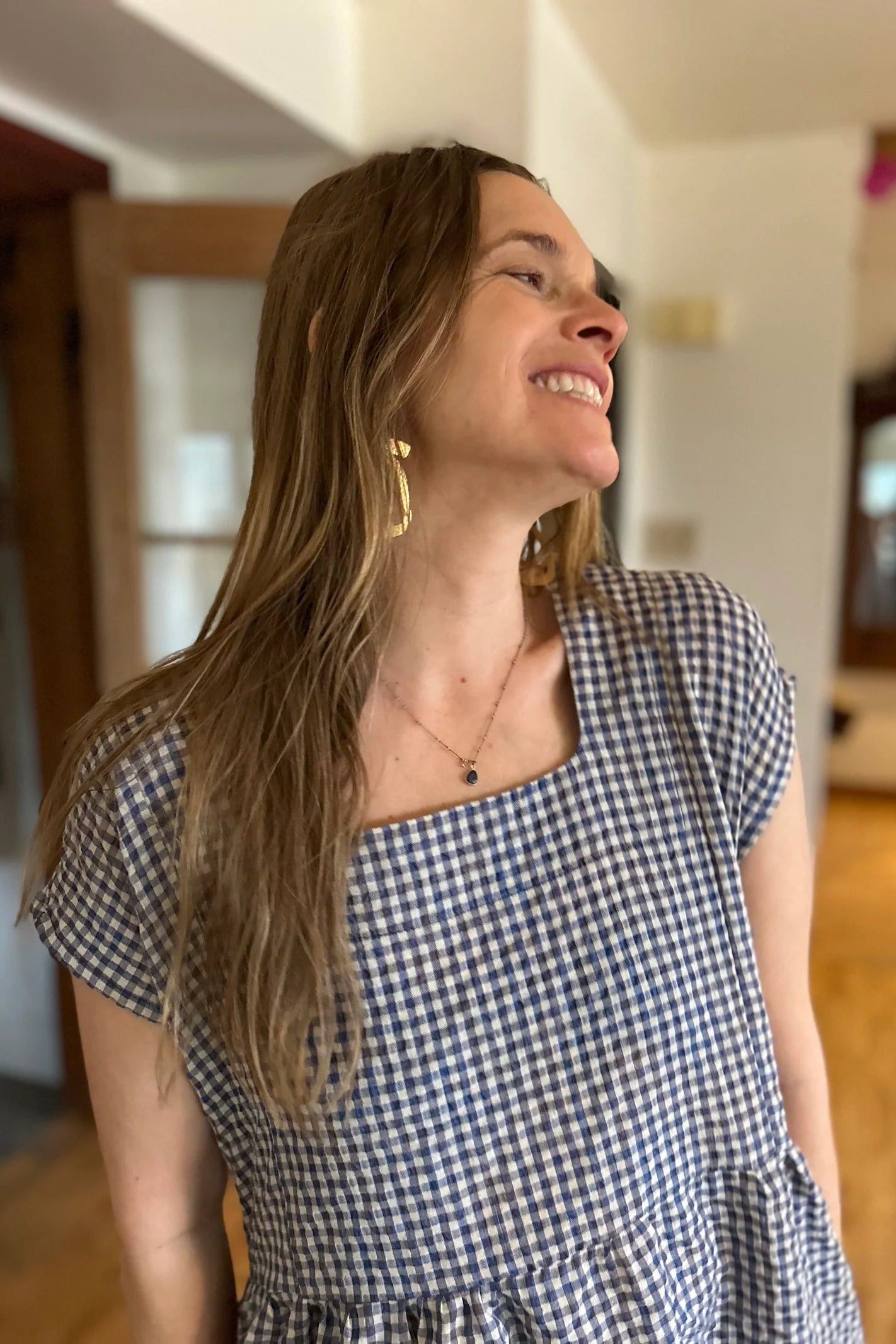 Close up view of a woman wearing the Lavendula Dress by Kazak in Sky Vichy/Gingham, a short sleeved square neck midi dress with three ruffles. She is standing in a room with wooden floors and a door.