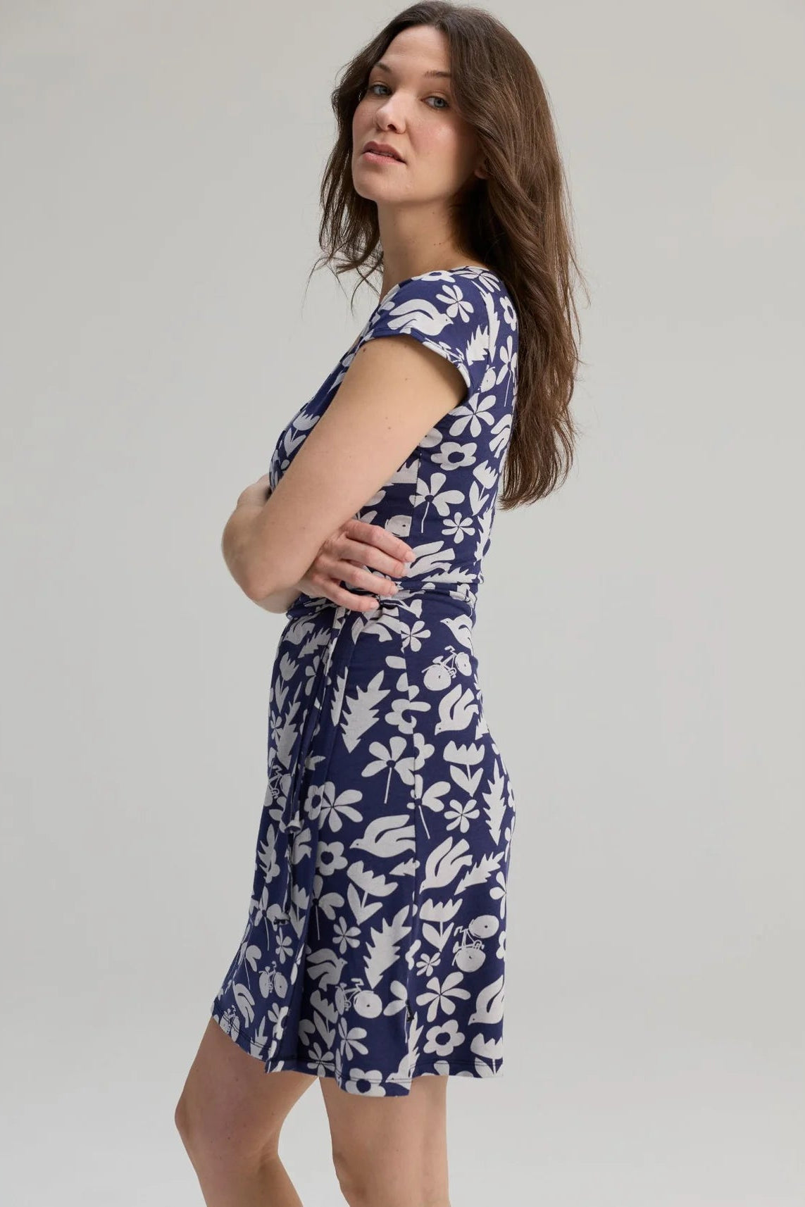 Side view of awoman wearing the Libellule Dress by Message Factory in Blue Print, a short sleeved wrap dress that falls to the knee. She is standing against a plain background. 