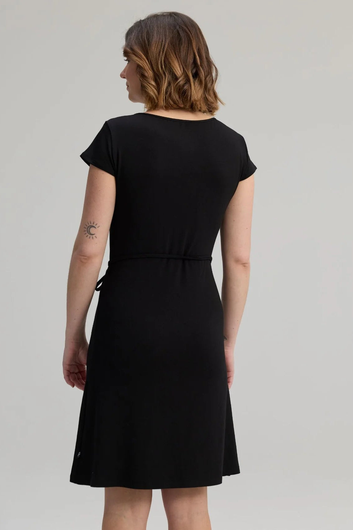 Back view of a woman wearing the Libellule Dress by Message Factory in Black, a short sleeved wrap dress that falls to the knee. She is standing against a plain background. 
