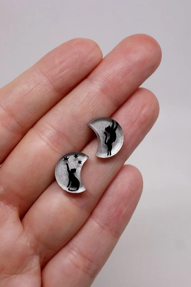 Silver moon shaped earrings with a black cat design held between fingers against a plain background/