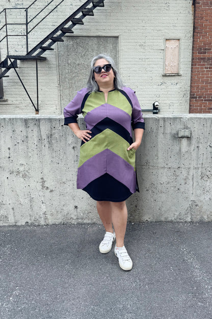A woman wearing the Lorraine Dress by Solomia in Avocado/Purple Navy, an A-line colour-blocked dress in stretch denim with 3/4 sleeves and a knee-length hem. She is standing outside in front of a cement wall. 