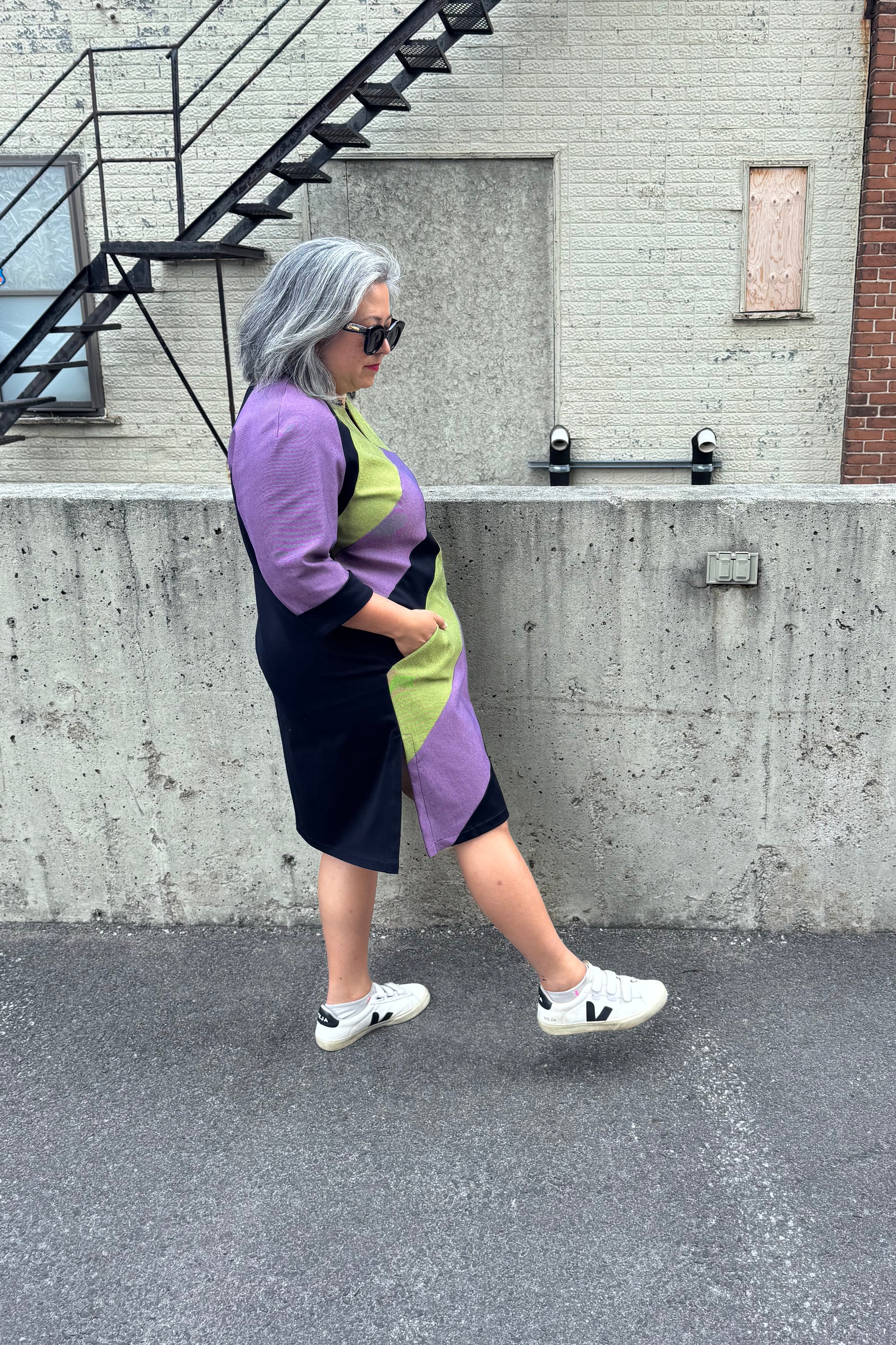 Side view of a woman wearing the Lorraine Dress by Solomia in Avocado/Purple Navy, an A-line colour-blocked dress in stretch denim with 3/4 sleeves and a knee-length hem. She is standing outside in front of a cement wall. 