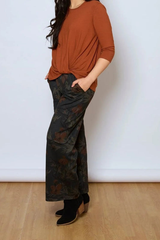A woman wearing the Luisa Pants by Tangente in Floral, featuring an elastic waist with pleats, straight legs, and side pockets. She is wearing them with a rust coloured top and standing in front of a white background. 