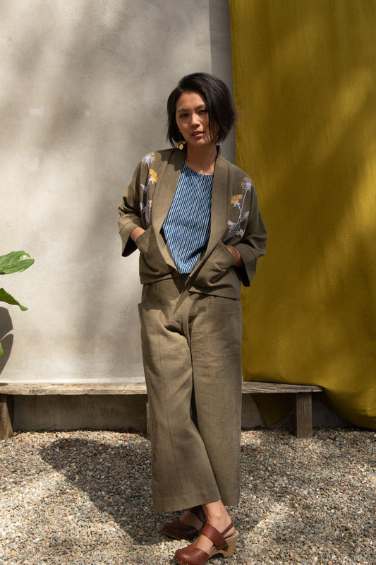 A woman wearing the Magnolia Screen-printed Jacket from Kazak in Olive with Dandelions, a short kimono style jacket with hand-screen printed dandelions, open front, 3/4-length sleeves, and big patch pockets. She is wearing it with matching pants and standing outside by a grey wall with a yellow curtain. 