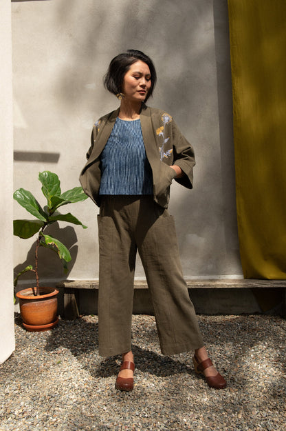 A woman wearing the Magnolia Screen-printed Jacket from Kazak in Olive with Dandelions, a short kimono style jacket with hand-screen printed dandelions, open front, 3/4-length sleeves, and big patch pockets. She is wearing it with matching pants and standing outside by a grey wall with a yellow curtain. 