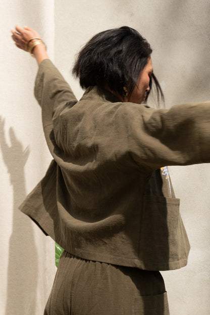 Back view of a woman wearing the Magnolia Screen-printed Jacket from Kazak in Olive with Dandelions, a short kimono style jacket with hand-screen printed dandelions, open front, 3/4-length sleeves, and big patch pockets. She is wearing it with matching pants and standing outside by a grey wall.