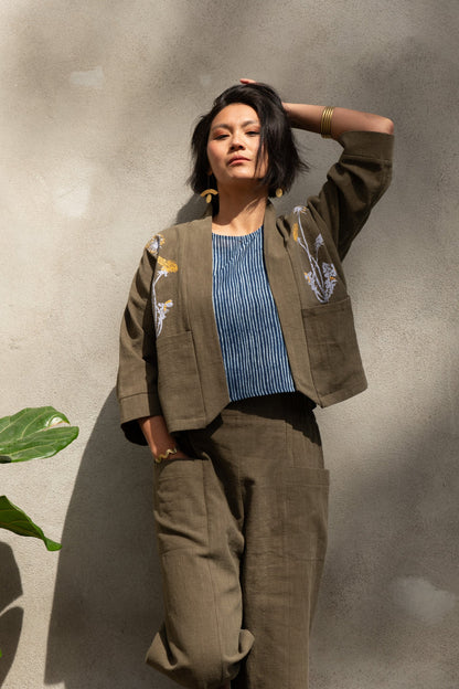 A woman wearing the Magnolia Screen-printed Jacket from Kazak in Olive with Dandelions, a short kimono style jacket with hand-screen printed dandelions, open front, 3/4-length sleeves, and big patch pockets. She is wearing it with matching pants and standing outside by a grey wall. 