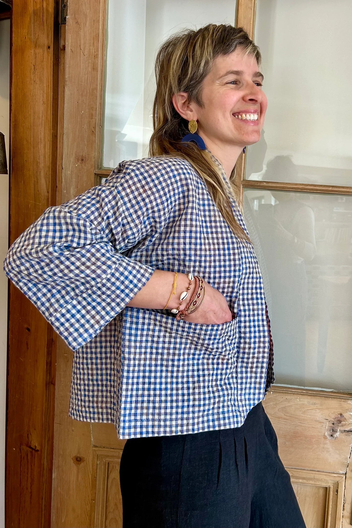 Side view of a woman wearing the Magnolia Jacket from Kazak in Sky Vichy/Gingham print, a kimono style jacket with 3/4 sleeves and large patch pockets. She is waering it with a red top and navy pants and standing in front of a door. 
