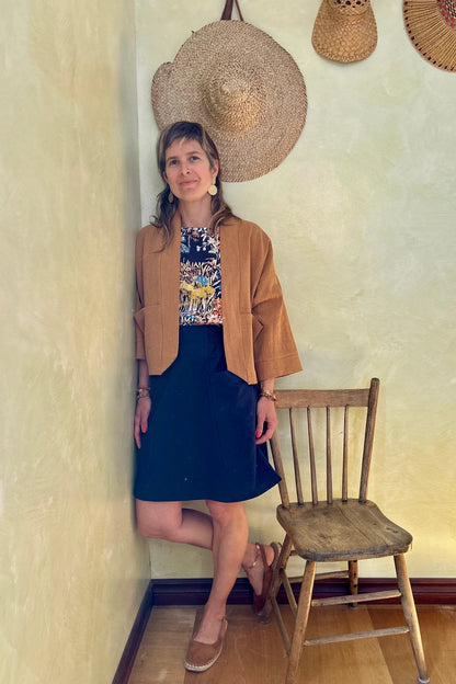 A woman wearing the Magnolia Jacket from Kazak in Turmeric, a kimono style jacket with 3/4 sleeves and large patch pockets. She is wearing it with a floral top navy and standing in a room with a wooden chair. 