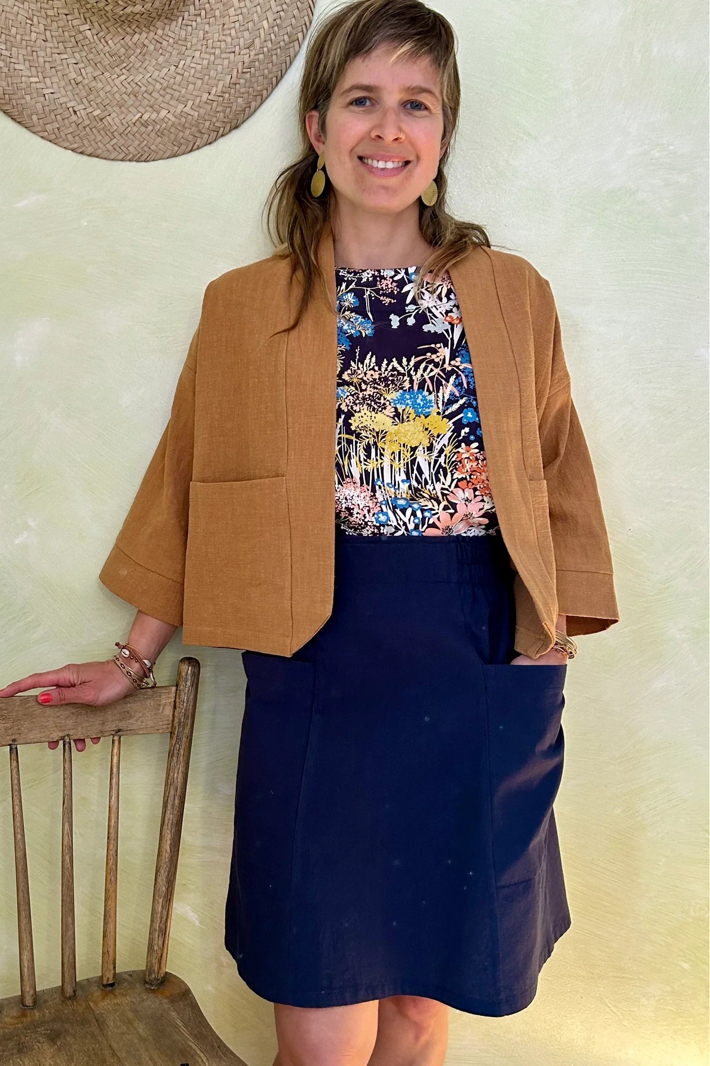 A woman wearing the Magnolia Jacket from Kazak in Turmeric, a kimono style jacket with 3/4 sleeves and large patch pockets. She is wearing it with a floral top navy and standing in a room with a wooden chair. 