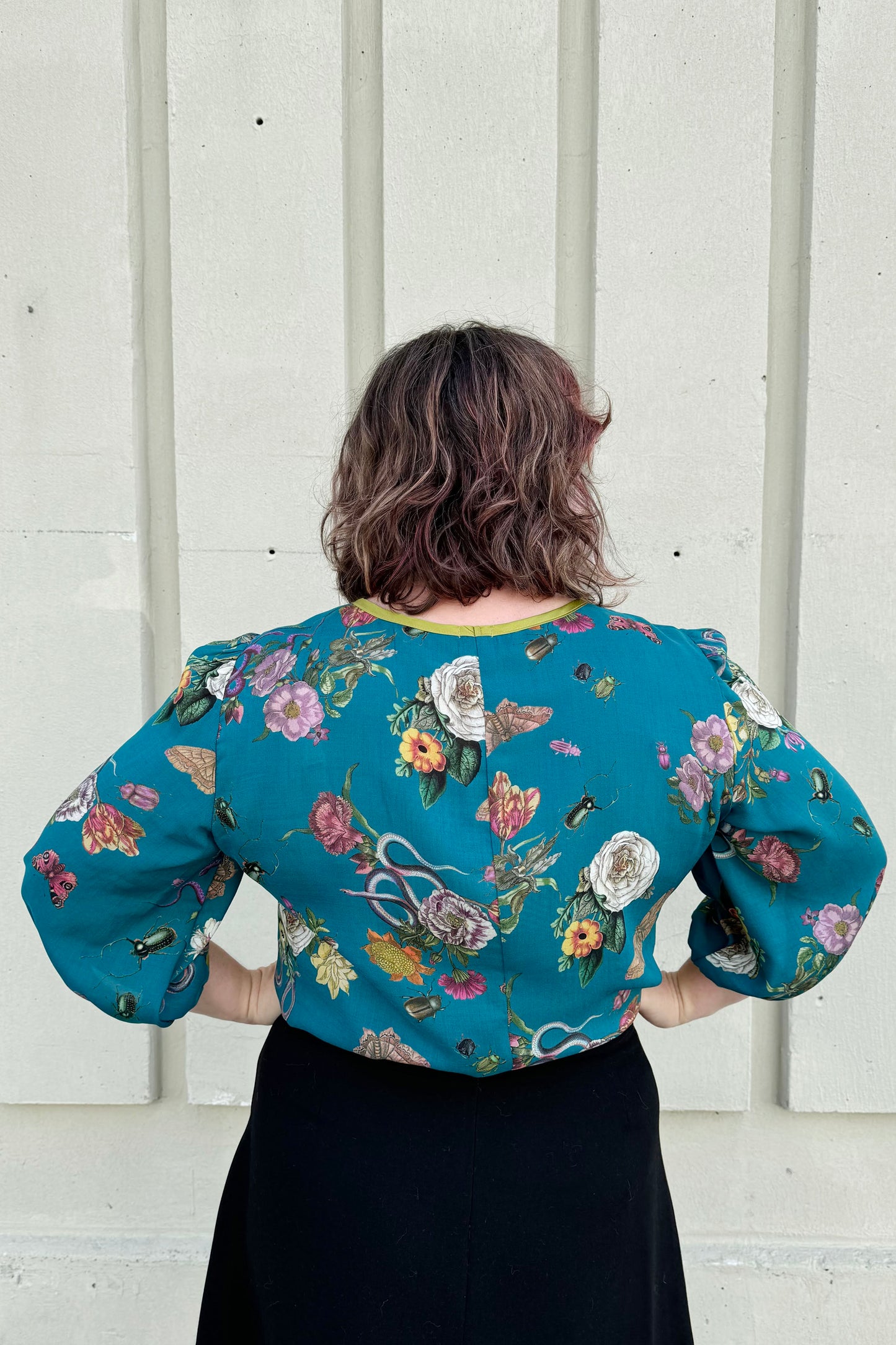 Back view of a woman wearing the Lydia Creeper Blouse by Mandala in Bayou. It features a vintage floral print with snakes and insects, with a jewel neck, puffed 3/4 sleeves, and a scooped hem. She is wearing a black skirt and standing in front of a beige wall. 