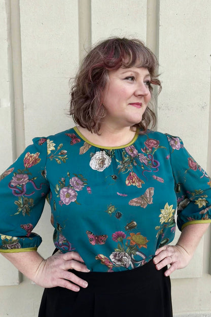 A woman wearing the Lydia Creeper Blouse by Mandala in Bayou. It features a vintage floral print with snakes and insects, with a jewel neck, puffed 3/4 sleeves, and a scooped hem. She is wearing a black skirt and standing in front of a beige wall. 