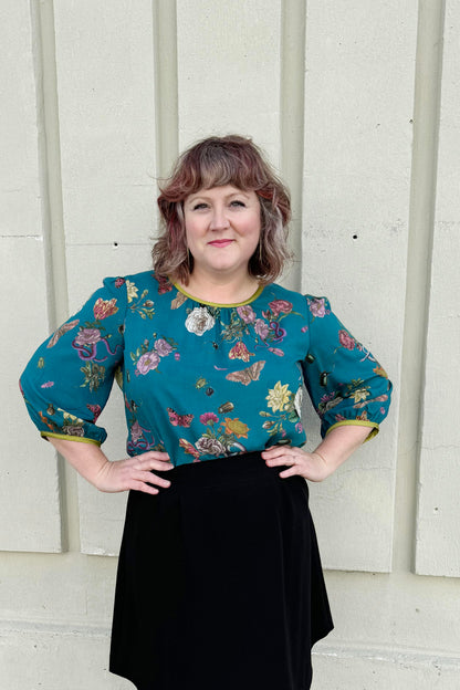 A woman wearing the Lydia Creeper Blouse by Mandala in Bayou. It features a vintage floral print with snakes and insects, with a jewel neck, puffed 3/4 sleeves, and a scooped hem. She is wearing a black skirt and standing in front of a beige wall. 