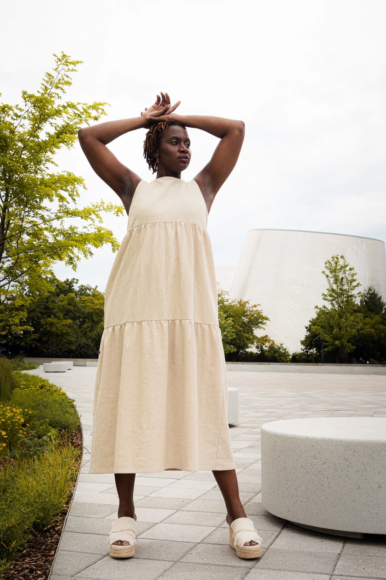 A woman wearing the Bari Dress by MAS in Oat, a tiered maxi dress with a sleeveless bodice and an open back with three ties. She is standing outdoors with trees and a building in the background.