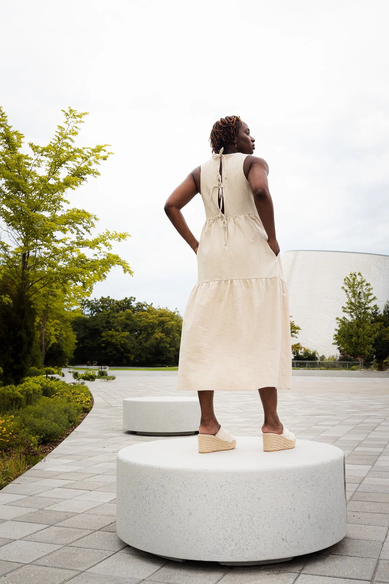 Back view of a woman wearing the Bari Dress by MAS in Oat, a tiered maxi dress with a sleeveless bodice and an open back with three ties. She is standing outdoors with trees and a building in the background.
