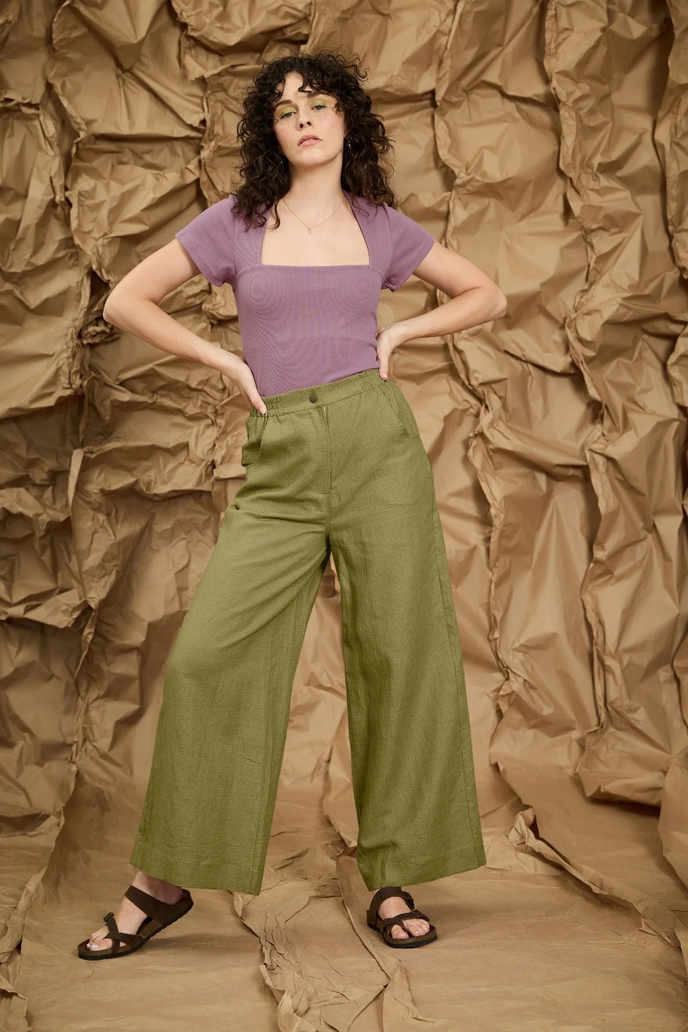A woman wearing the Matisse Trousers by Melow in Pesto check, full length wide leg pants with a zipper front and a high waist with elastic at the back and side and back patch pockets. She is wearing them with a purple top and standing in front of a textured brown backdrop