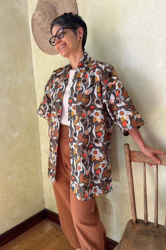 A woman wearing the Mirano Jacket by Kazak in Olive Floral, a mid-thigh length open kimono jacket with 3/4 sleeves. She is wearing it with the Oxalis pants in Ginger and standing in a room by a wooden chair. 