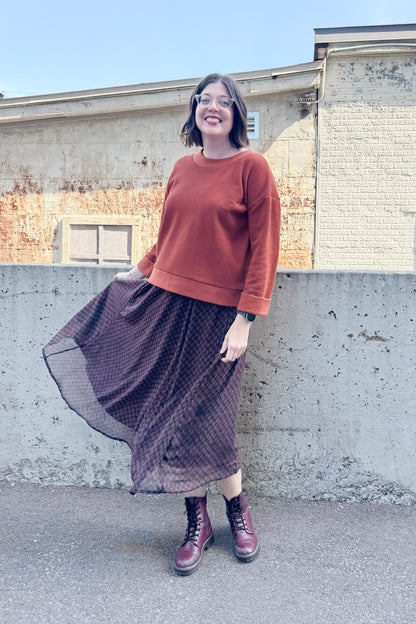 A woman wearing the Espresso Skirt by Cherry Bobin in Black and Brown print, a long and flowing chiffon skirt that's fully lined. She's wearing it with the Mocha Sweater in Bronze and standing outside in front of a brick building.
