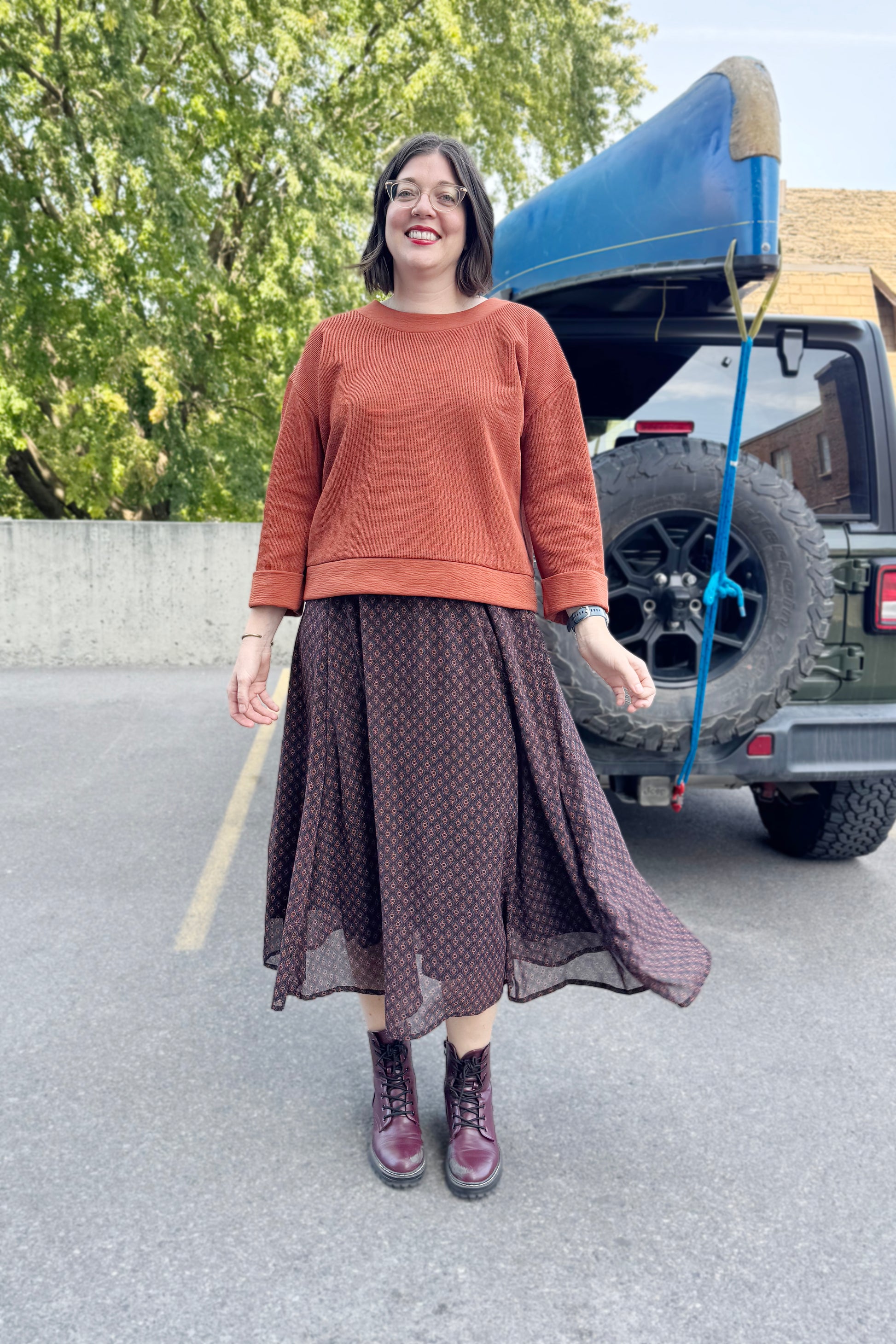 A woman wearing the Espresso Skirt by Cherry Bobin in Black and Brown print with the Mocha Sweater in Bronze and standing outside in front of a vehicle . 