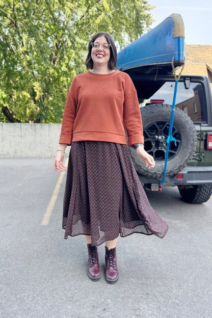 A woman wearing the Espresso Skirt by Cherry Bobin in Black and Brown print with the Mocha Sweater in Bronze and standing outside in front of a vehicle . 
