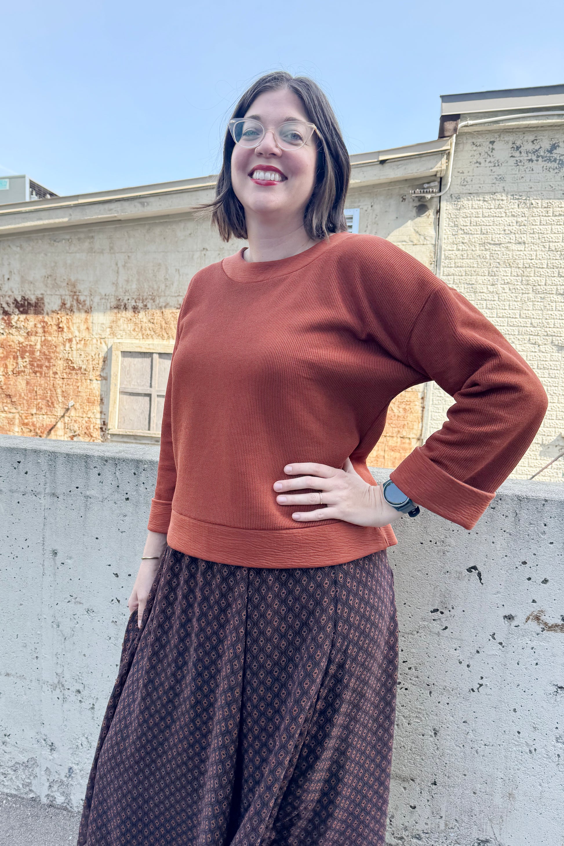 A woman wearing the Espresso Skirt by Cherry Bobin in Black and Brown print with the Mocha Sweater in Bronze and standing outside in front of a brick wall.  