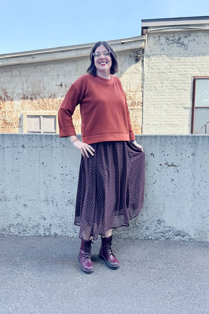A woman wearing the Espresso Skirt by Cherry Bobin in Black and Brown print, a long and flowing chiffon skirt that's fully lined. She's wearing it with the Mocha Sweater in Bronze and standing outside in front of a brick building.
