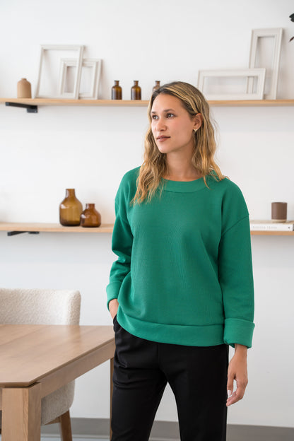 A woman wearing the Mocha SWeater by Cherry Bobin  in Green, with a unique knit, fold-over sleeves, and a relaxed fit. She is standing in a room with shelves and a table.