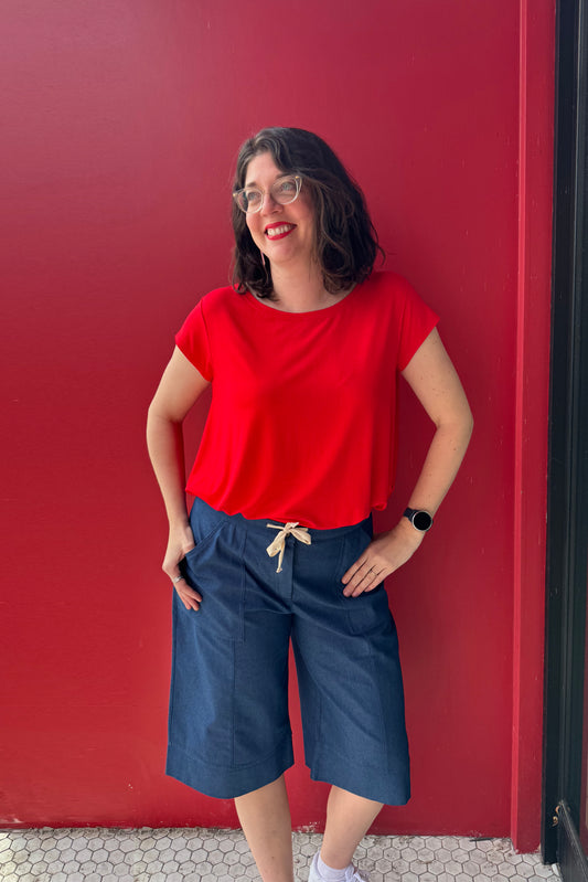 A woman wearing the Brooke Capri by Julei, wide-legged denim capris with a white drawstring and elastic at the back waist. She is wearing them with  a red shirt and standing against a red wall.