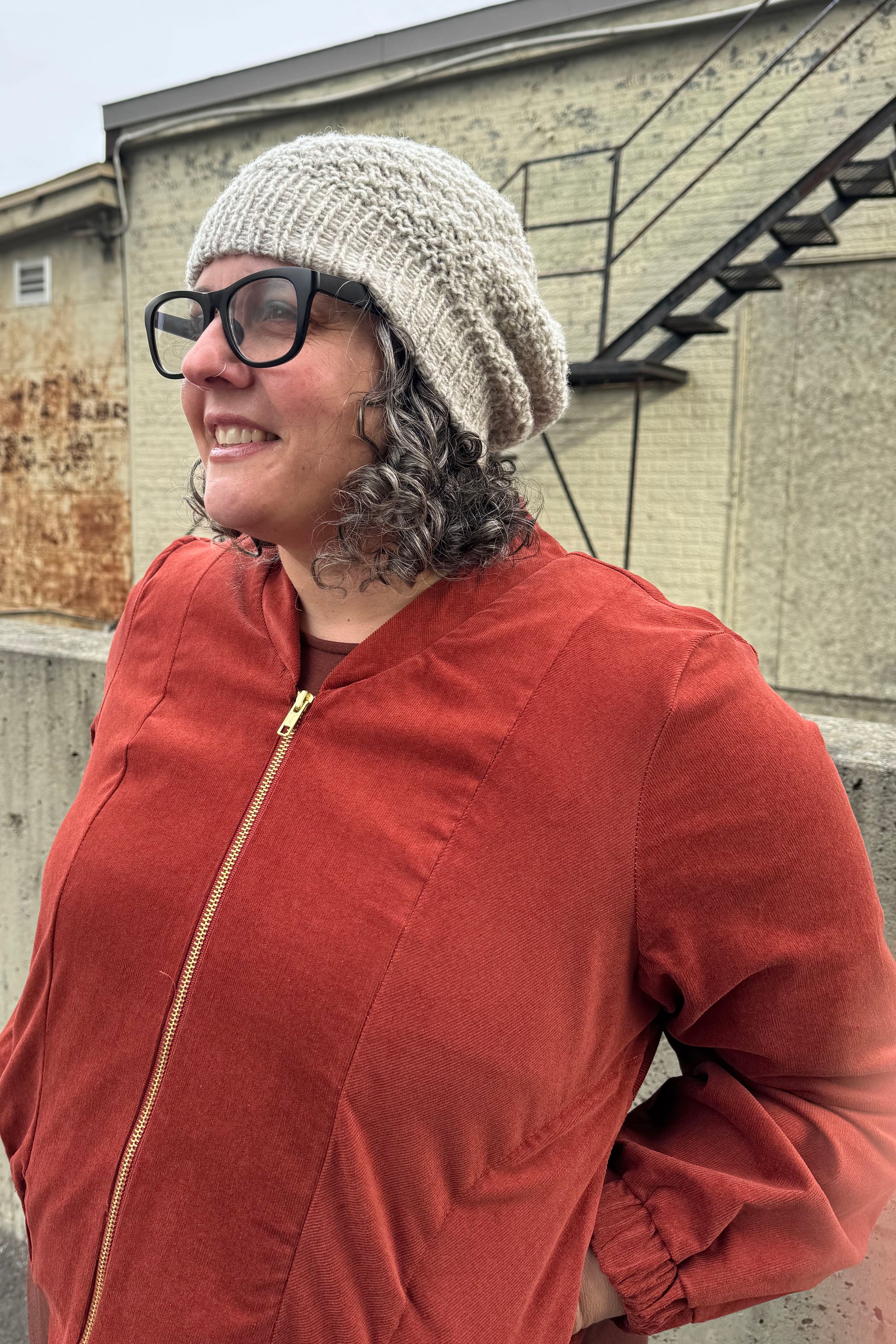 A woman wearing the Beret by Charlevoix Pure Laine in Grey with the Redrawn Zip Jacket by Eve Lavoie in Terracotta Corduroy. She is standing outside. 