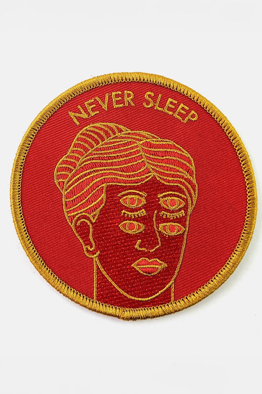 The Never Sleep Patch from Badaboöm Studio, circular embroidered patch with a red background, gold border, and a stylized face with two sets of open eyes. The words 'NEVER SLEEP' are embroidered in gold at the top.